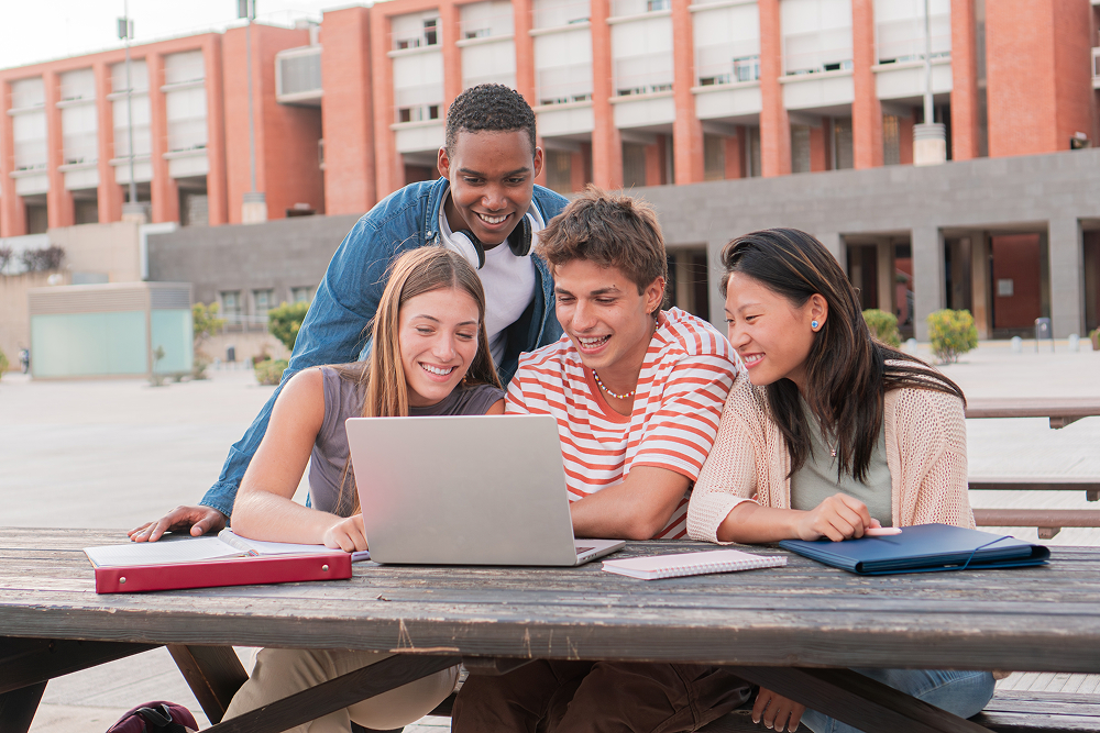 4 students standing around laptop on college campus. 